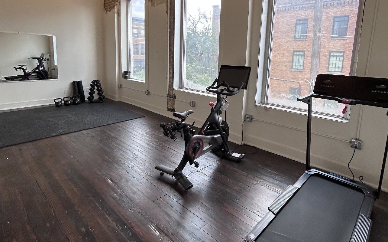 A gym room with a stationary bike, a treadmill, kettlebells, and dumbbells. Large windows with a city view and a mirror on the wall.