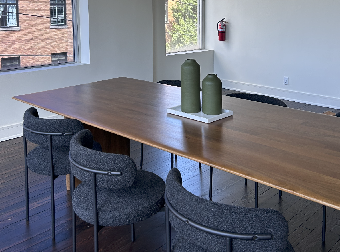 A coworking space with a large wooden table with four black fabric chairs around it, two green vases on a white tray in the center, large windows and hardwood floors.
