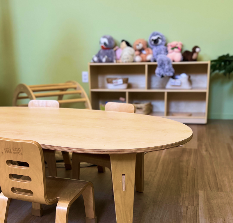 A wooden activity table with chairs in a playroom, with a bookshelf filled with plush toys in the background.