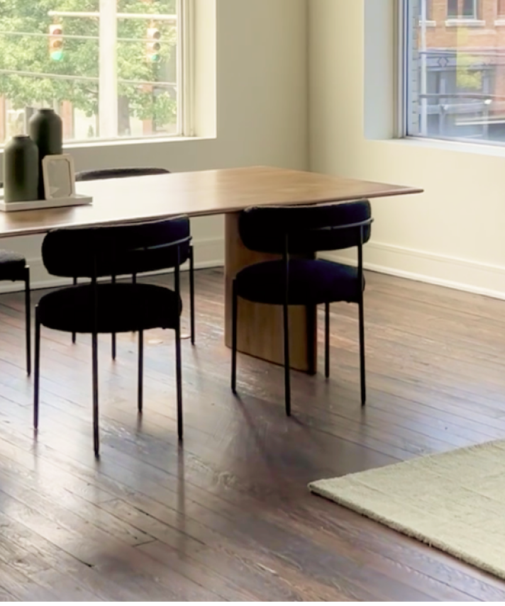 Modern dining area with a wooden table, two black chairs, a small rug, and large windows letting in natural light.