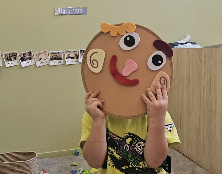 Child holding a craft mask in front of their face, resembling a smiling face with large eyes, curly cheeks, and a tongue sticking out, in a classroom with a green wall.