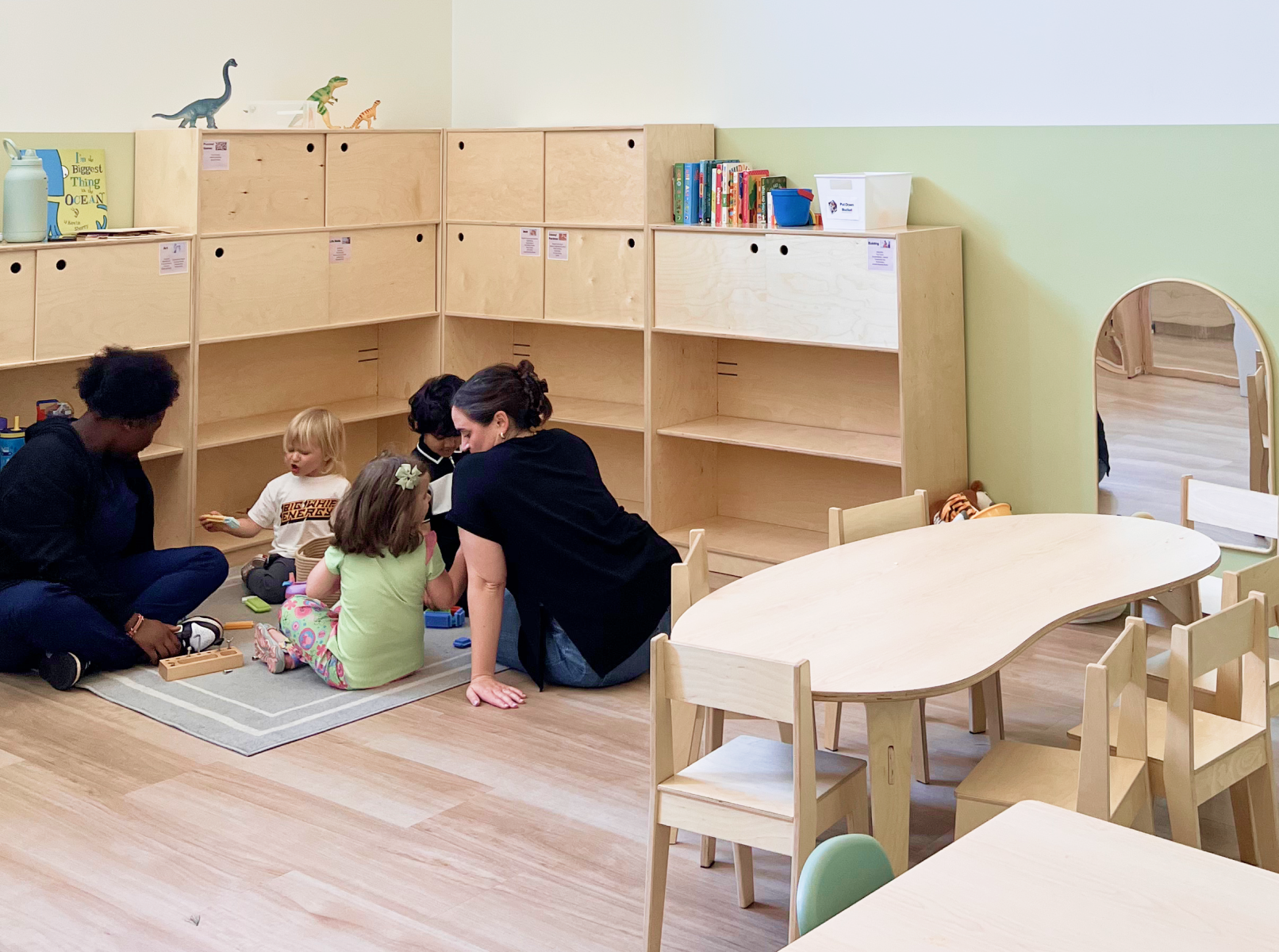 Photo showing a group of kids and their teachers playing in the corner of a classroom, surrounded by shelves and toys.