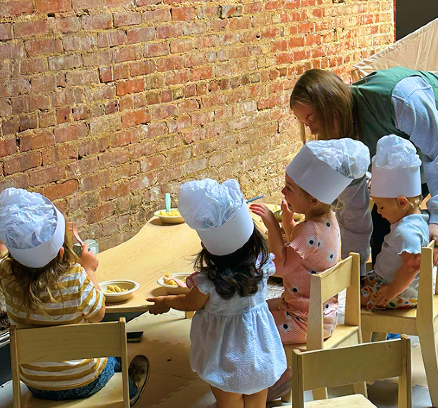 Children dressed as chefs with white chef hats. Sitting at a wooden table at Moxi with bowls of snacks.