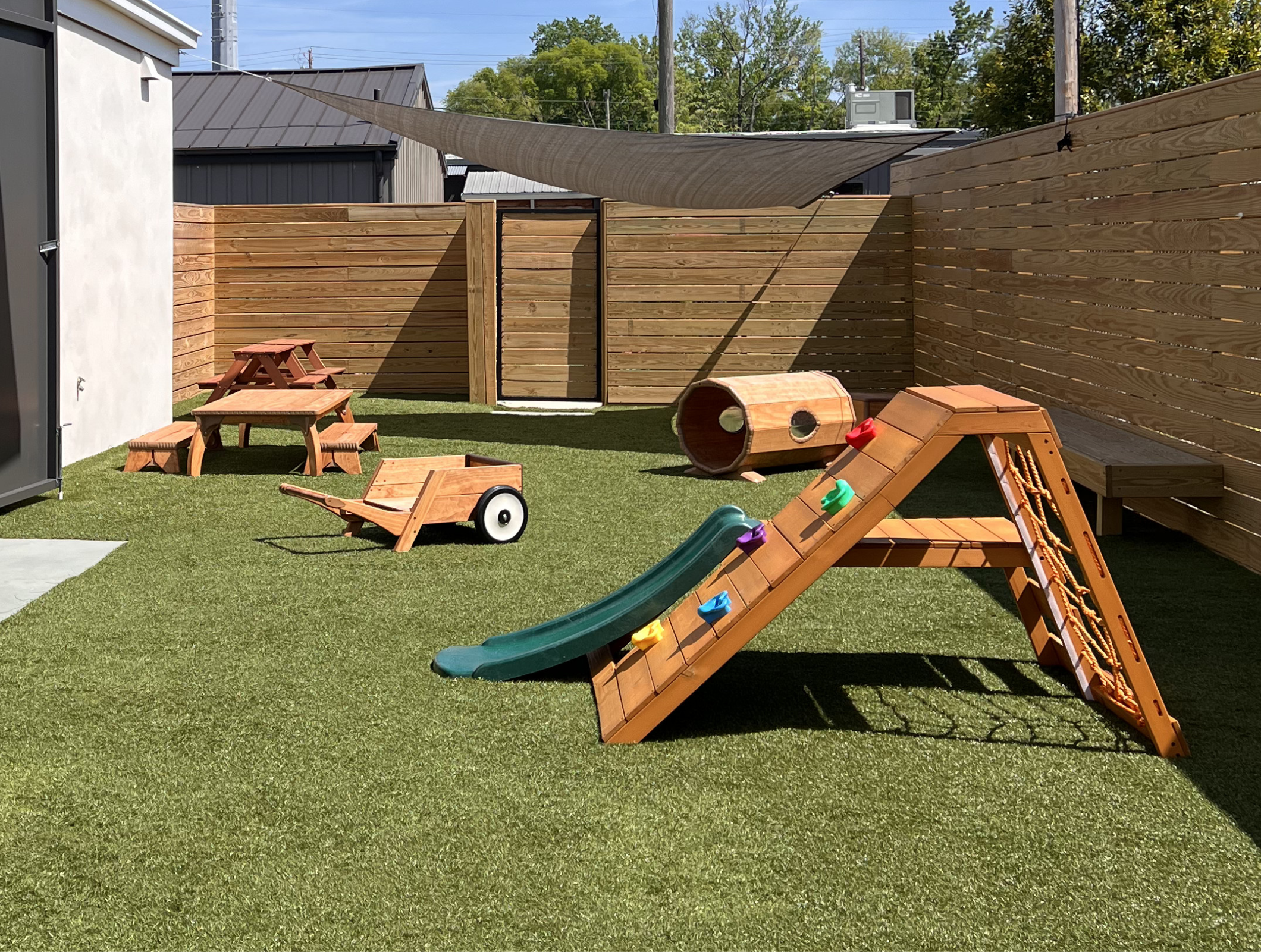 Backyard with wooden fence, children's playground equipment including slide and tunnel, small picnic table, and artificial grass, with trees and buildings in the background.