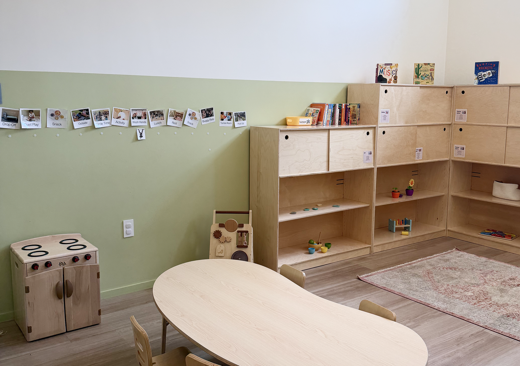 A classroom with light green and white walls, wooden shelves filled with books, toys, and plants, and a round table with chairs.