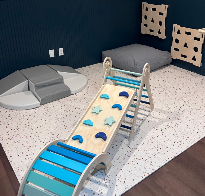 Children's play area with a wooden slide, cushioned mat, bed, and sensory panels on the wall.