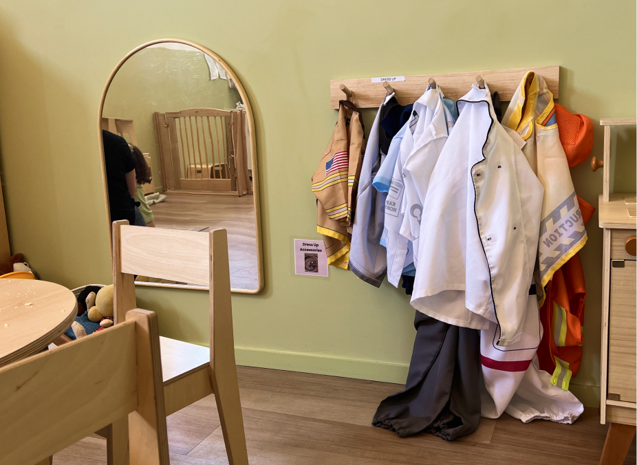 A corner of a room with a wooden wall rack. A small sign reads 'Dress Up Accessories.' A round mirror on the wall reflects part of the room and a person with long hair. There are wood floors and a table with chairs in the room.