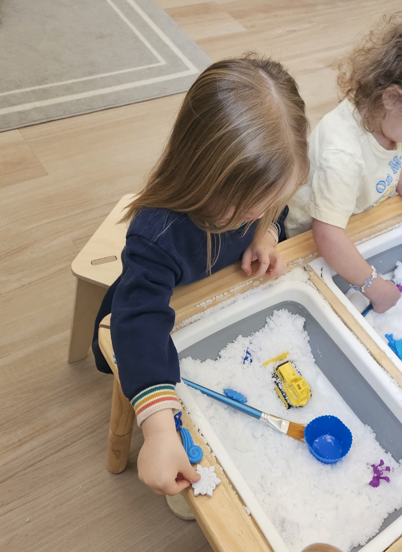 Child playing in a sensory bin filled with sand, along with small toy construction vehicles and tools.