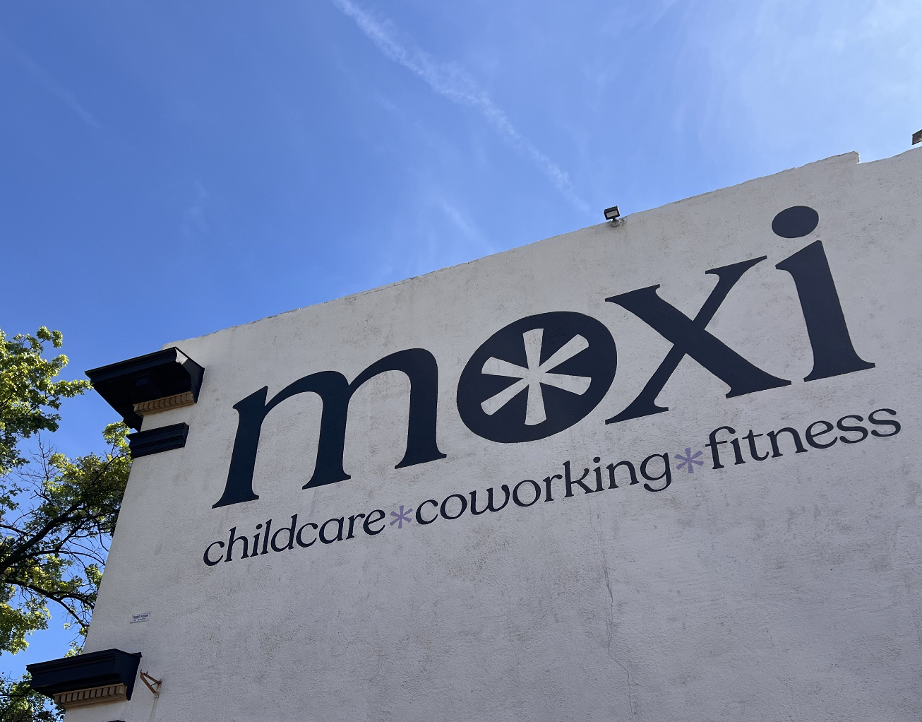 Exterior of the Moxi building with the large sign 'moxi' and the words 'childcare,' 'coworking,' and 'fitness' beneath it. There is a blue sky in the background.