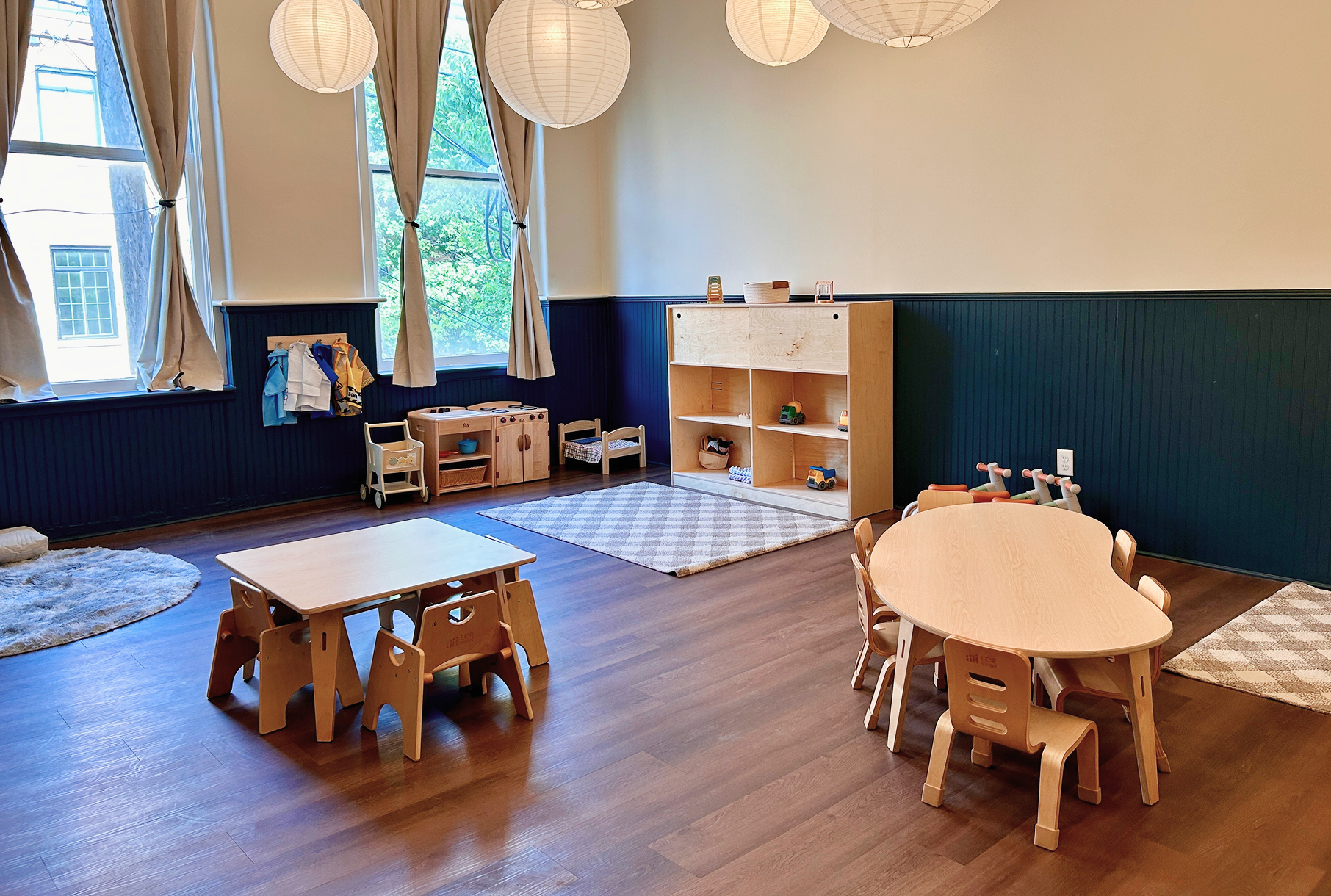A spacious and well-lit montessori children's playroom with wooden furniture, toy storage, small tables and chairs and large windows, decorated with round paper lanterns hanging from the ceiling.