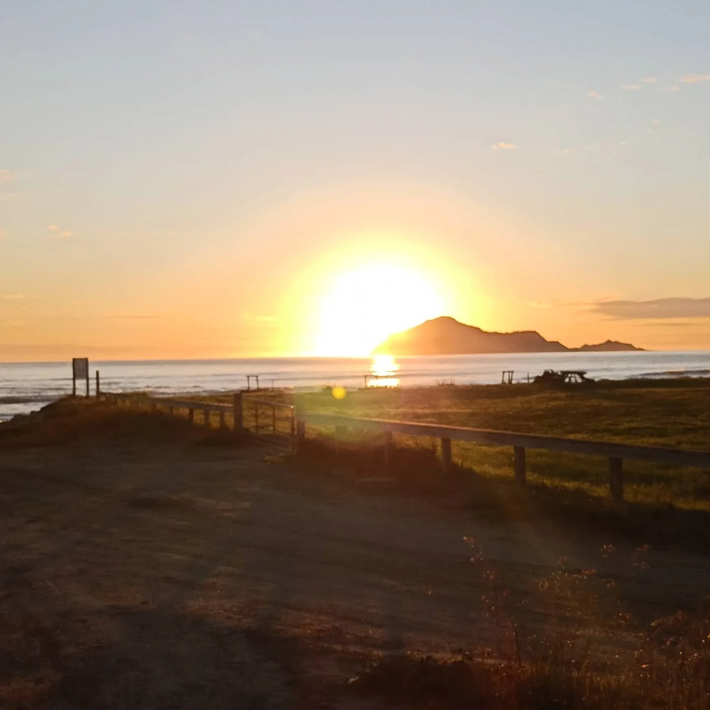 Sunset over the ocean with a mountain island in the distance, grassy area and fence in foreground.