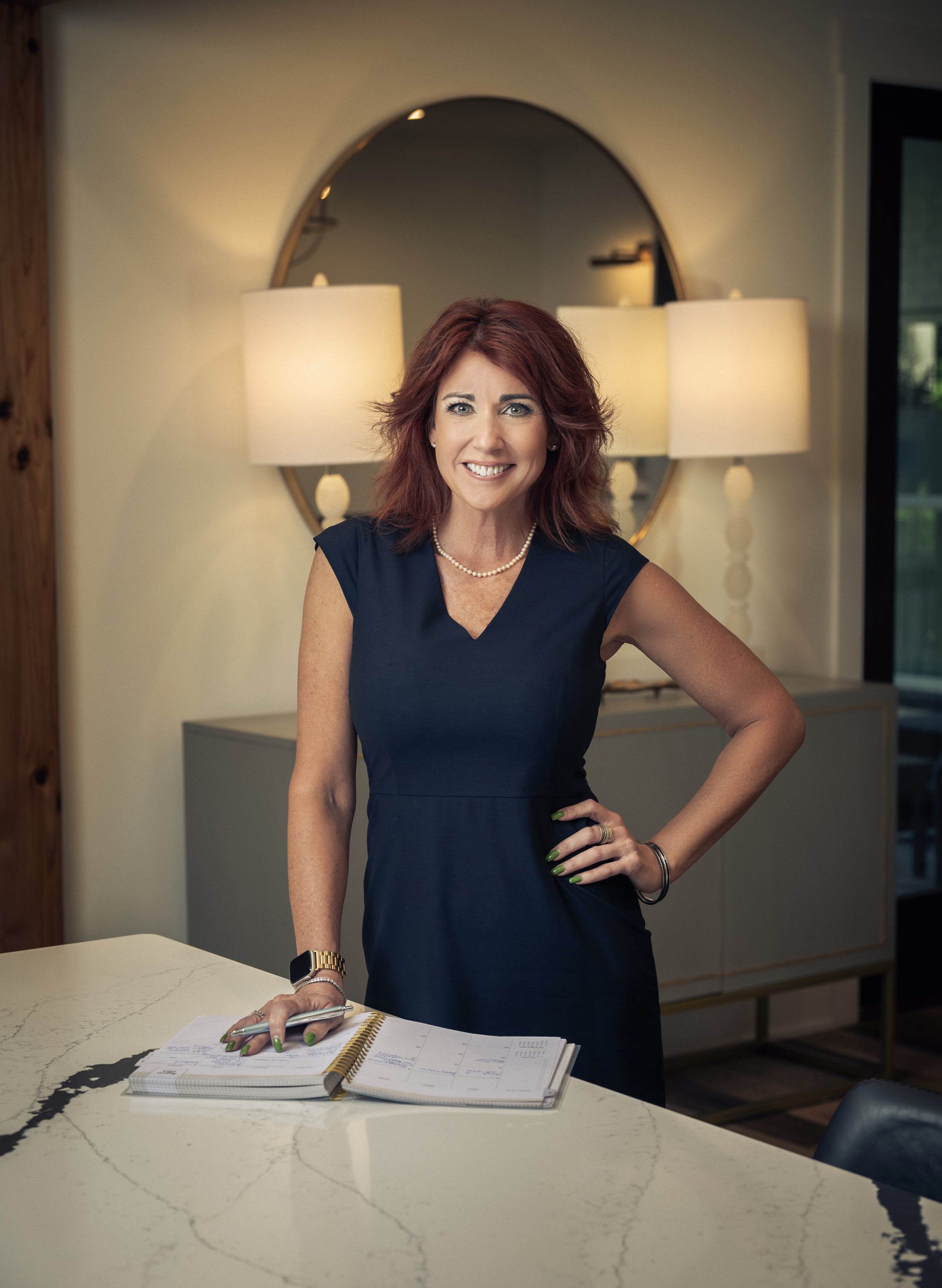 A woman with red hair, wearing a navy blue dress with a pearl necklace, standing in a modern room with a white marble table, a large mirror, and two lamps in the background, smiling at the camera.