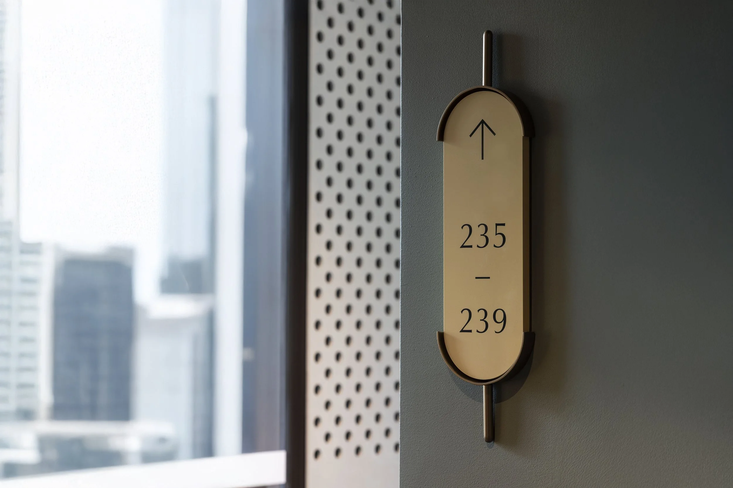 Close-up of a beige elevator directory sign with black arrow pointing up, room numbers 235 and 239, mounted on a wall near a window with natural light.