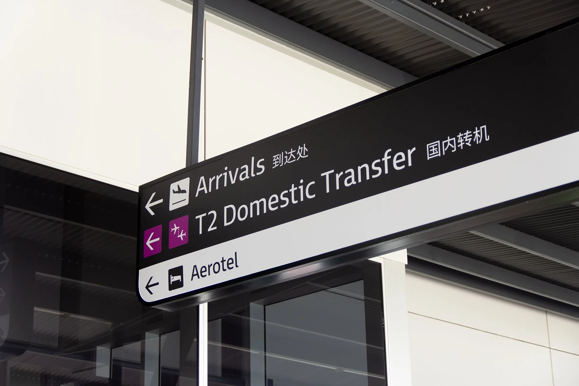 Sign at an airport showing directions to arrivals, domestic transfer, and Aerotel. The sign is black with white text and symbols.