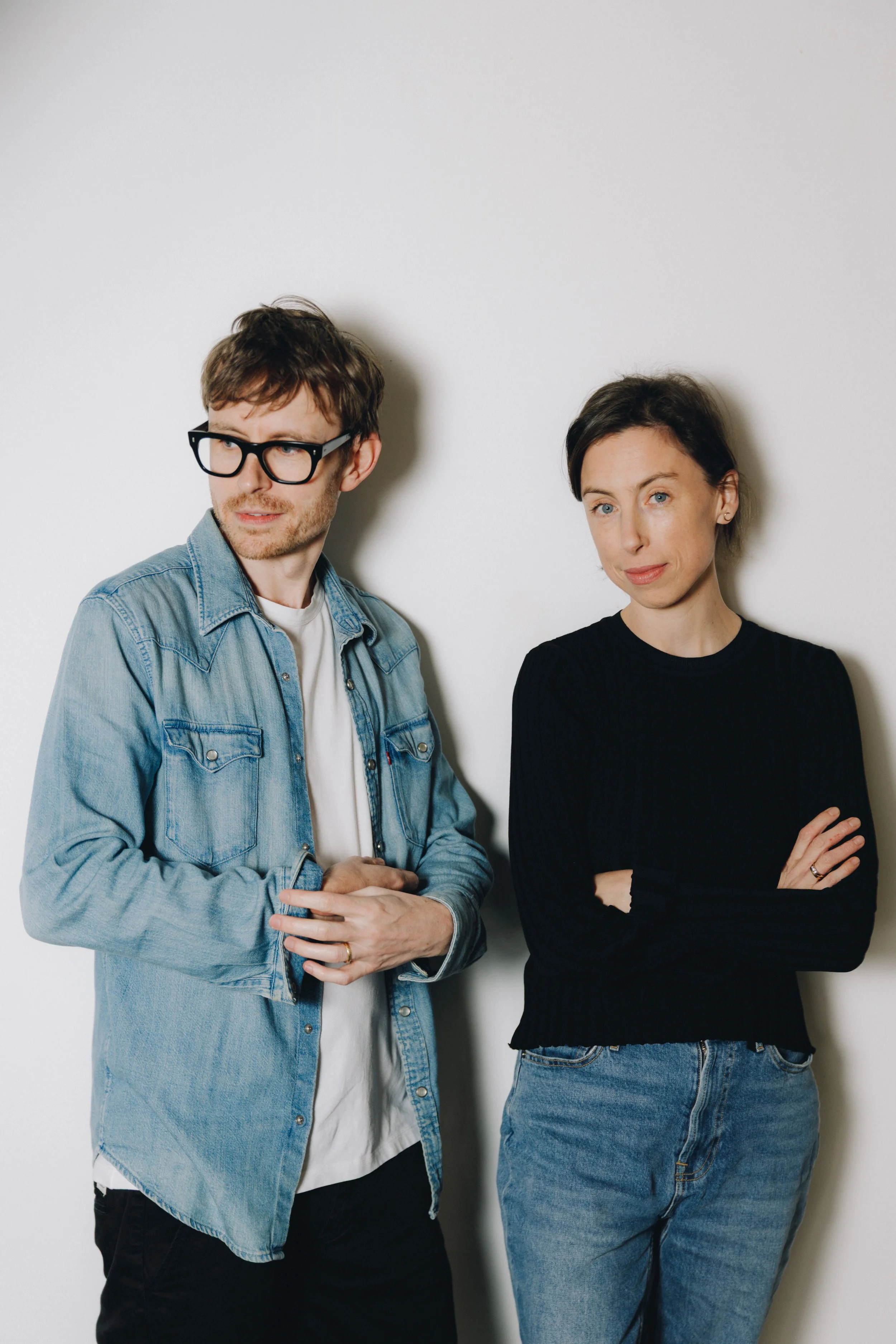 A man with glasses and a woman with short dark hair standing against a plain white wall.