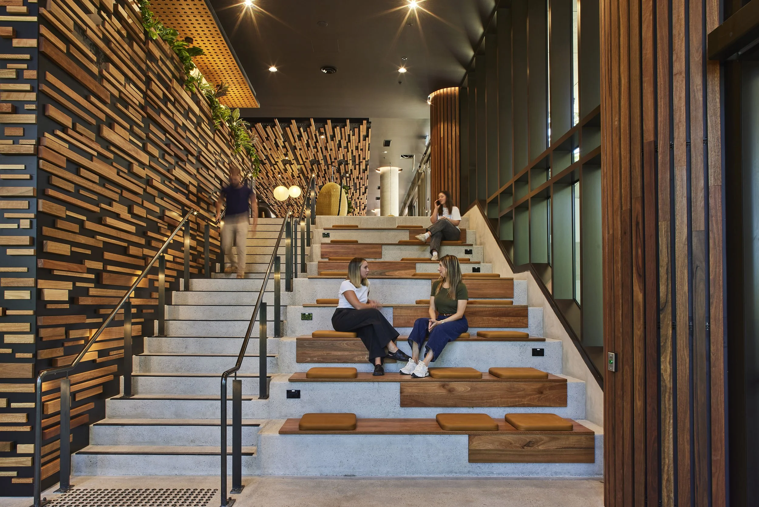 Interior of a modern building with wooden accents and a staircase with seating. People are sitting and walking, with one woman on the stairs talking on her phone. The space features decorative wood paneling, greenery, and warm lighting.