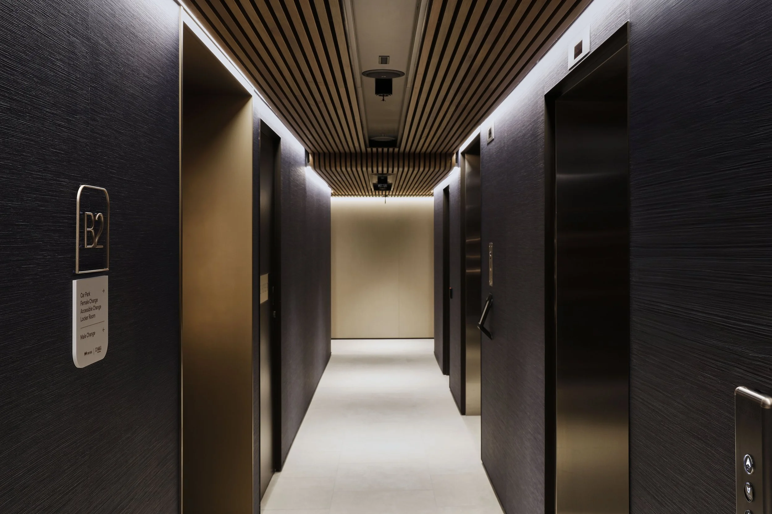 Dark modern lift lobby with timber ceiling battens, textured walls and wall-mounted wayfinding signage for Basement Level 2.
