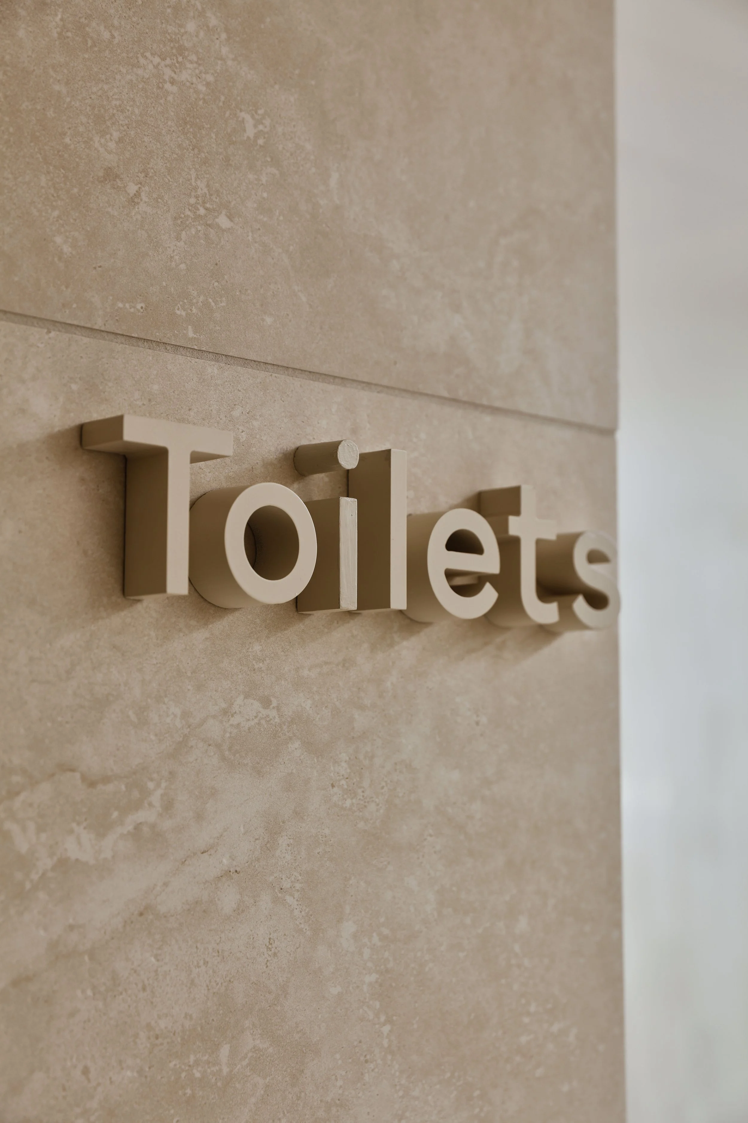 Close-up view of beige and light brown textured wall with small white decorative letters spelling 'Toilets' mounted on the wall.