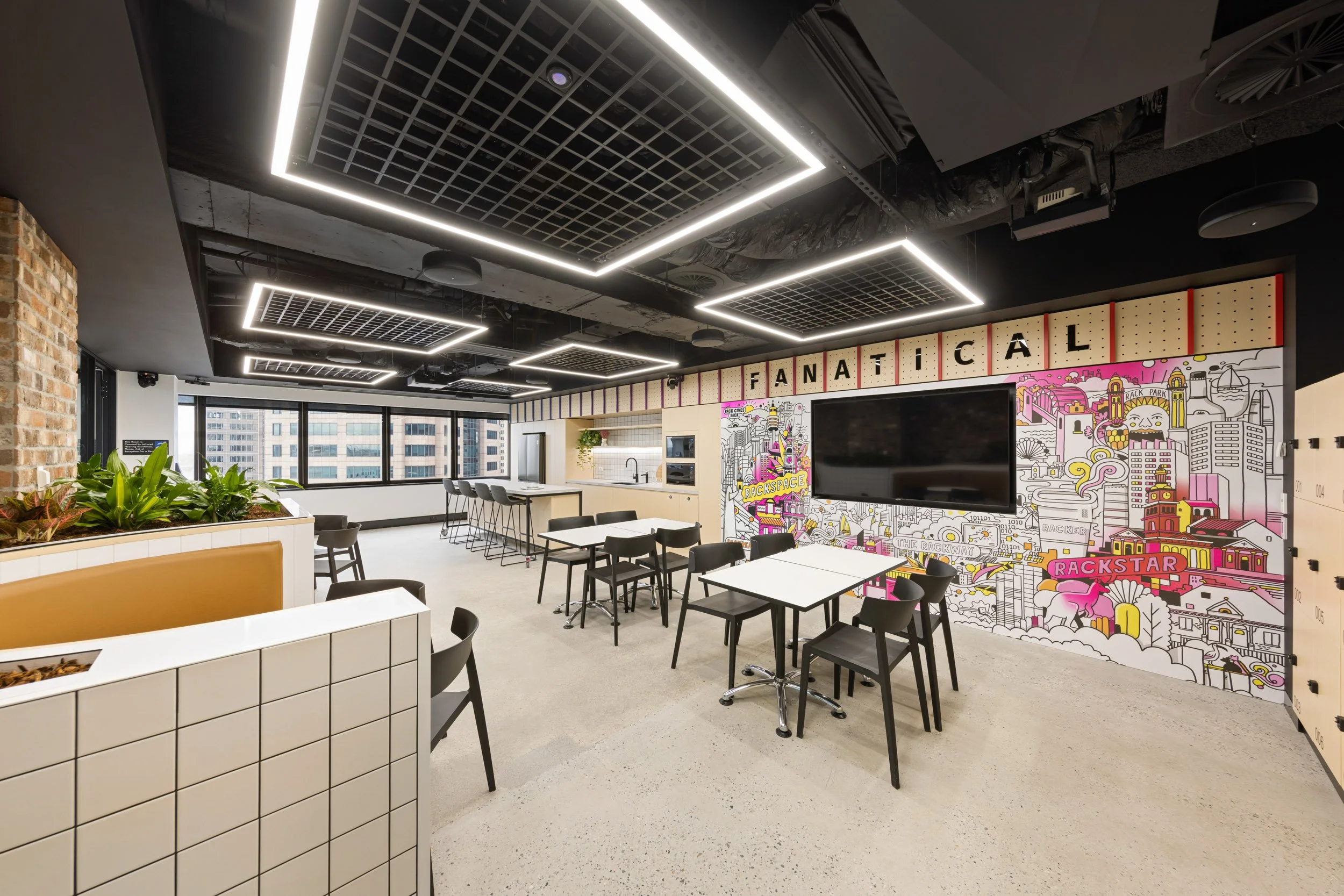 Modern office space with suspended ceiling panels, large windows, and a wall decorated with colorful urban cityscape murals. There are tables and chairs arranged for meetings or collaboration, along with potted plants adding greenery.