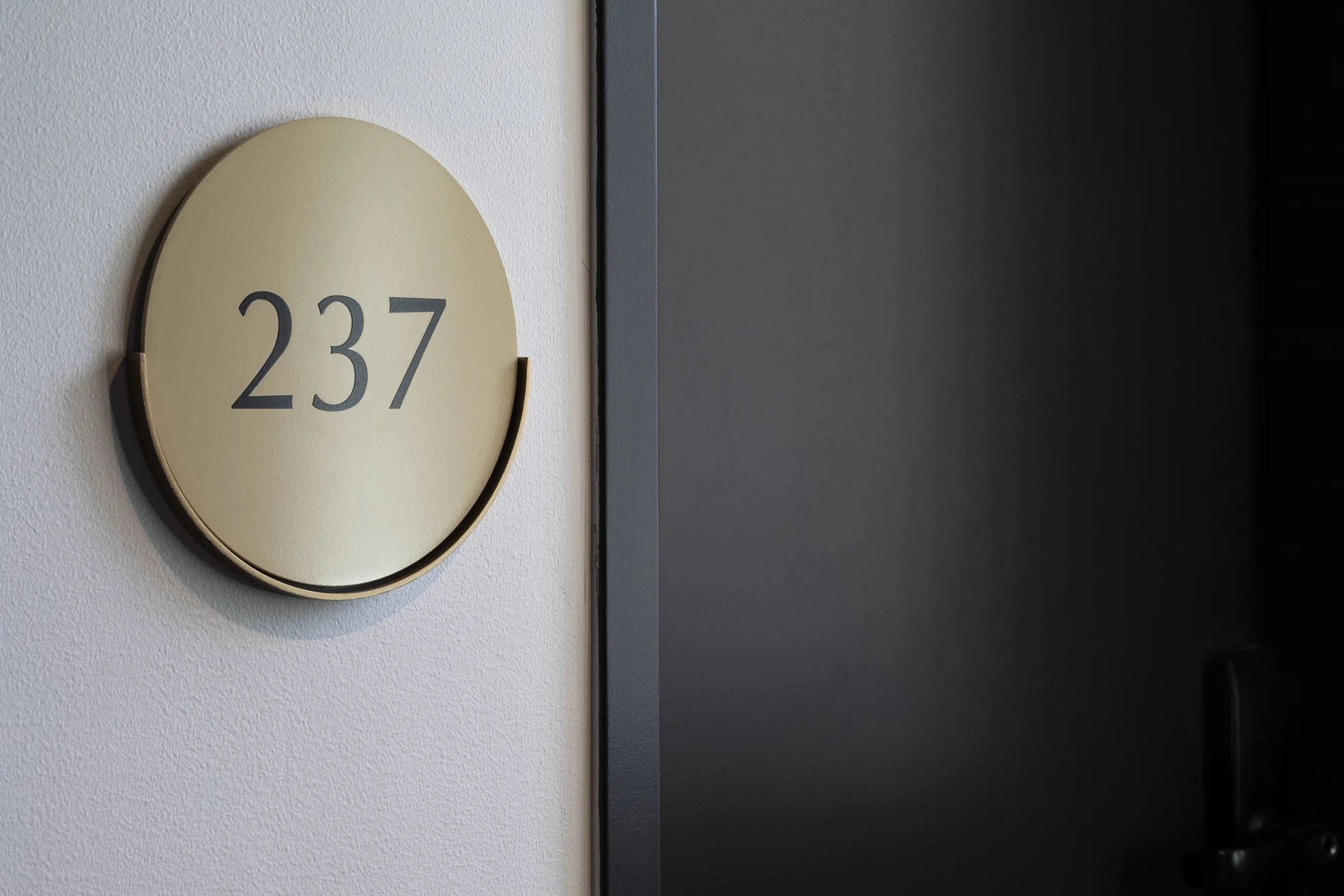 Close-up of a wall-mounted circular beige sign with the number 237 in black, situated on a white textured wall next to a dark gray surface.