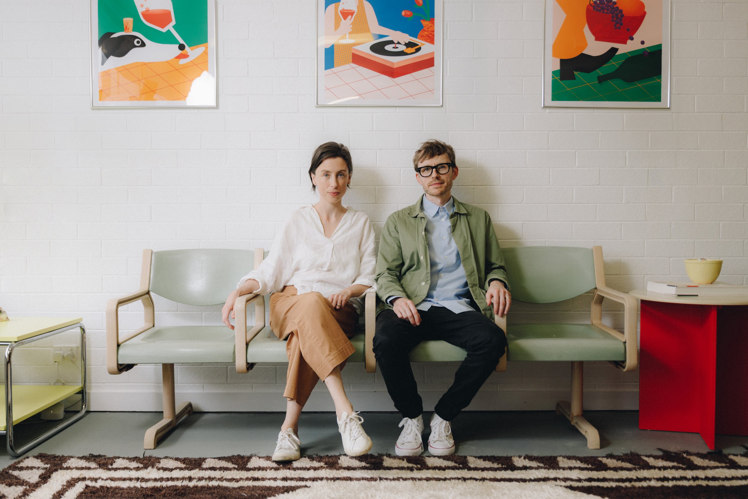 Two people sitting on a waiting room bench against a white brick wall with colorful abstract art. The woman has short dark hair, wearing a white top and tan pants, and the man has short brown hair, wearing glasses, a green jacket, and black pants. A small yellow bowl and a stack of books are on a side table nearby. A patterned brown and white carpet lies on the floor.