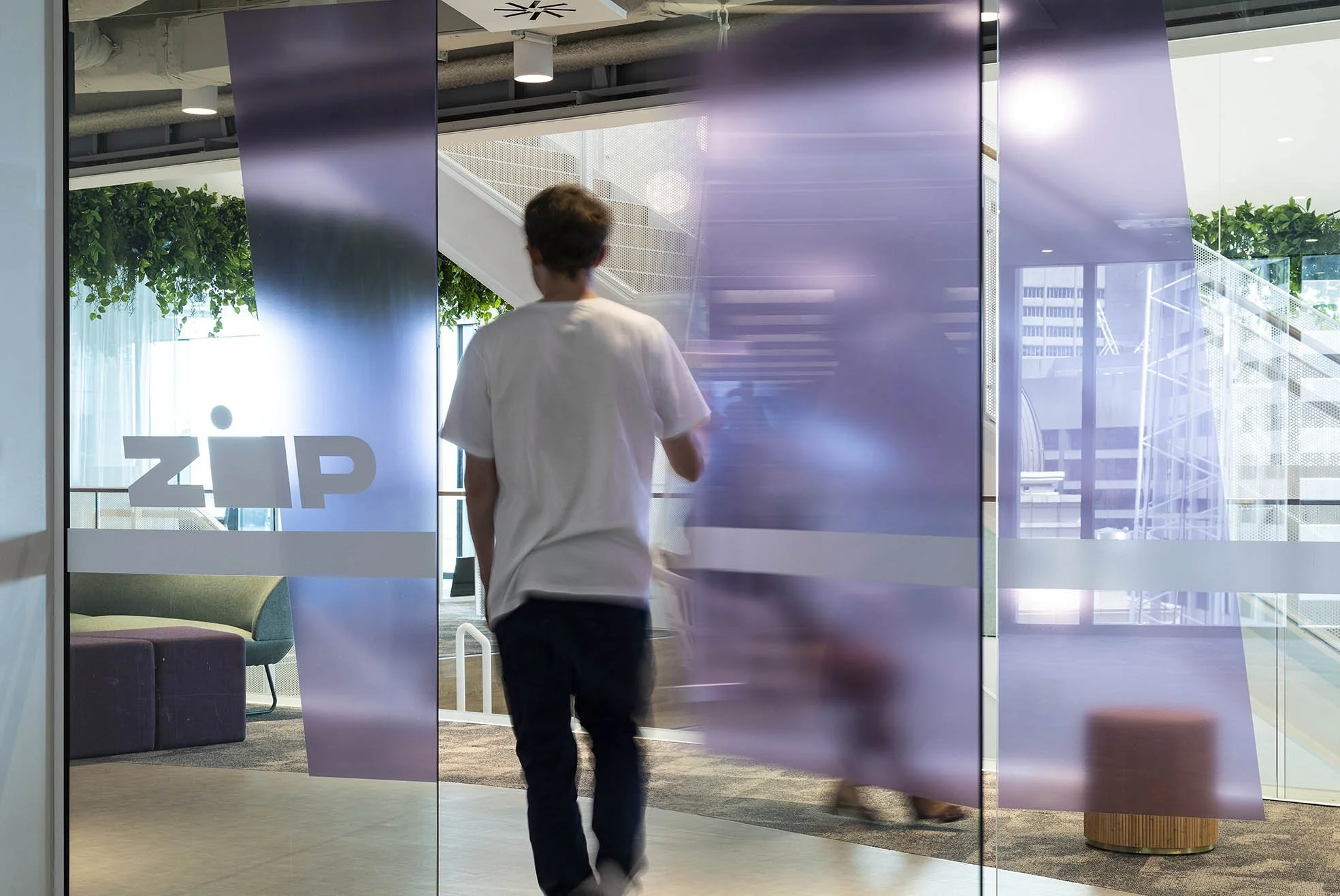A person in a white shirt walking into or out of a modern office space through glass doors with the word ‘ZIP’ on them. The office features bright natural light, green plants, colorful chairs, and a staircase in the background.