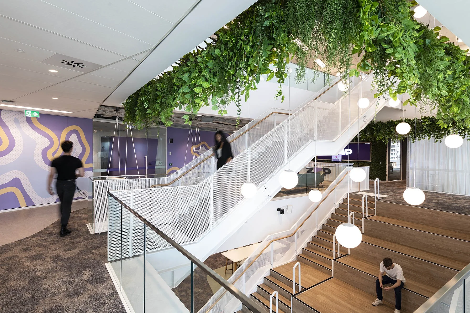 Modern interior of an office or commercial space with a green plant wall, staircase, hanging globe lights, and people walking and sitting.