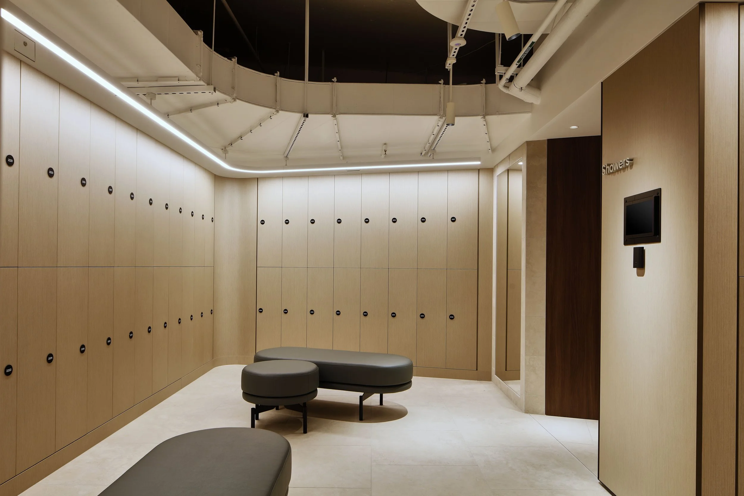 A modern locker room with beige lockers, black round seats, and wood paneling. Overhead lighting and ceiling fixtures are visible.