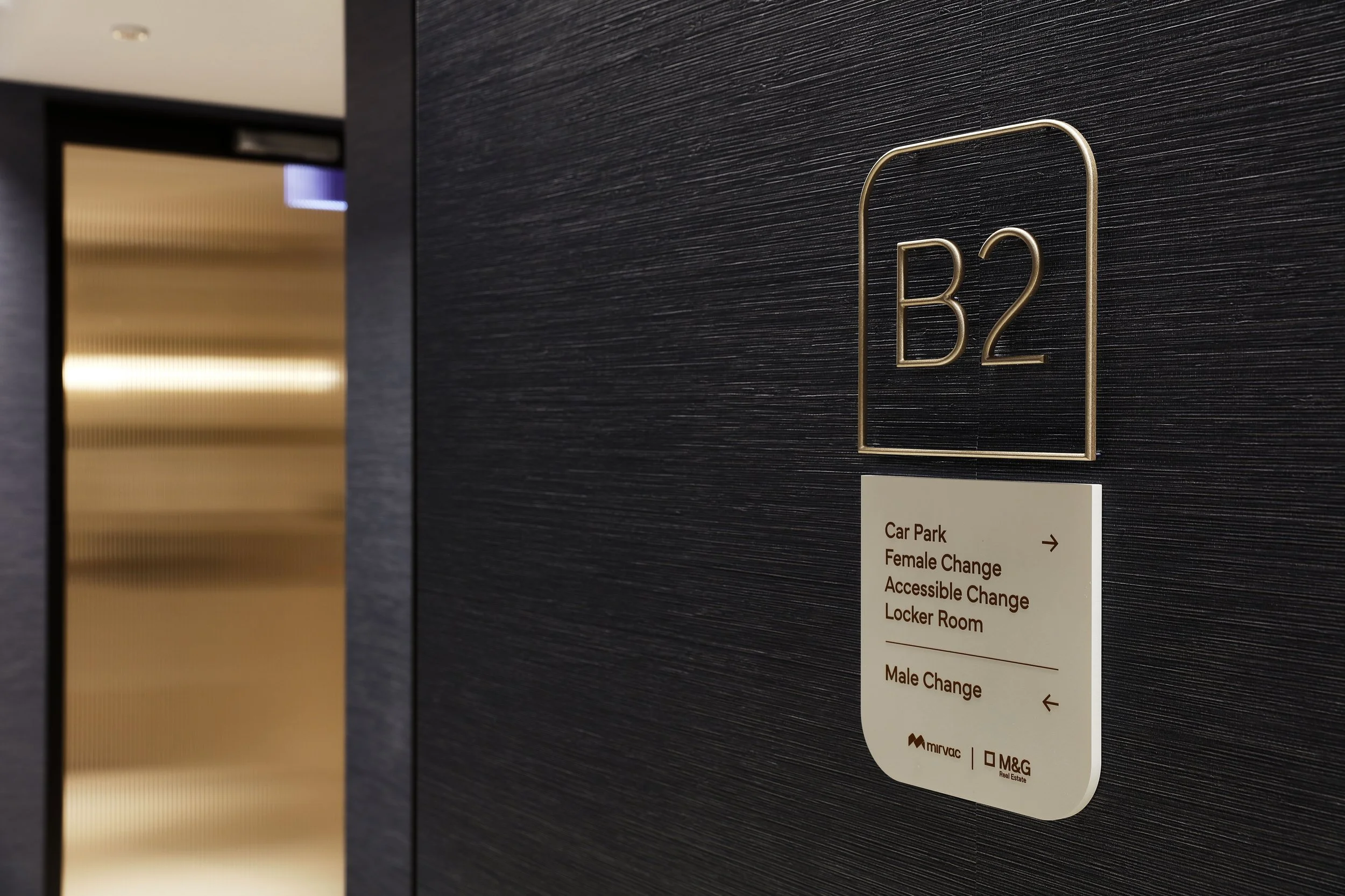 A dark wall with a gold-colored sign reading 'B2' and a white directory sign listing services such as car park, female change, accessible change, locker room, and male change, located near an elevator with golden doors.