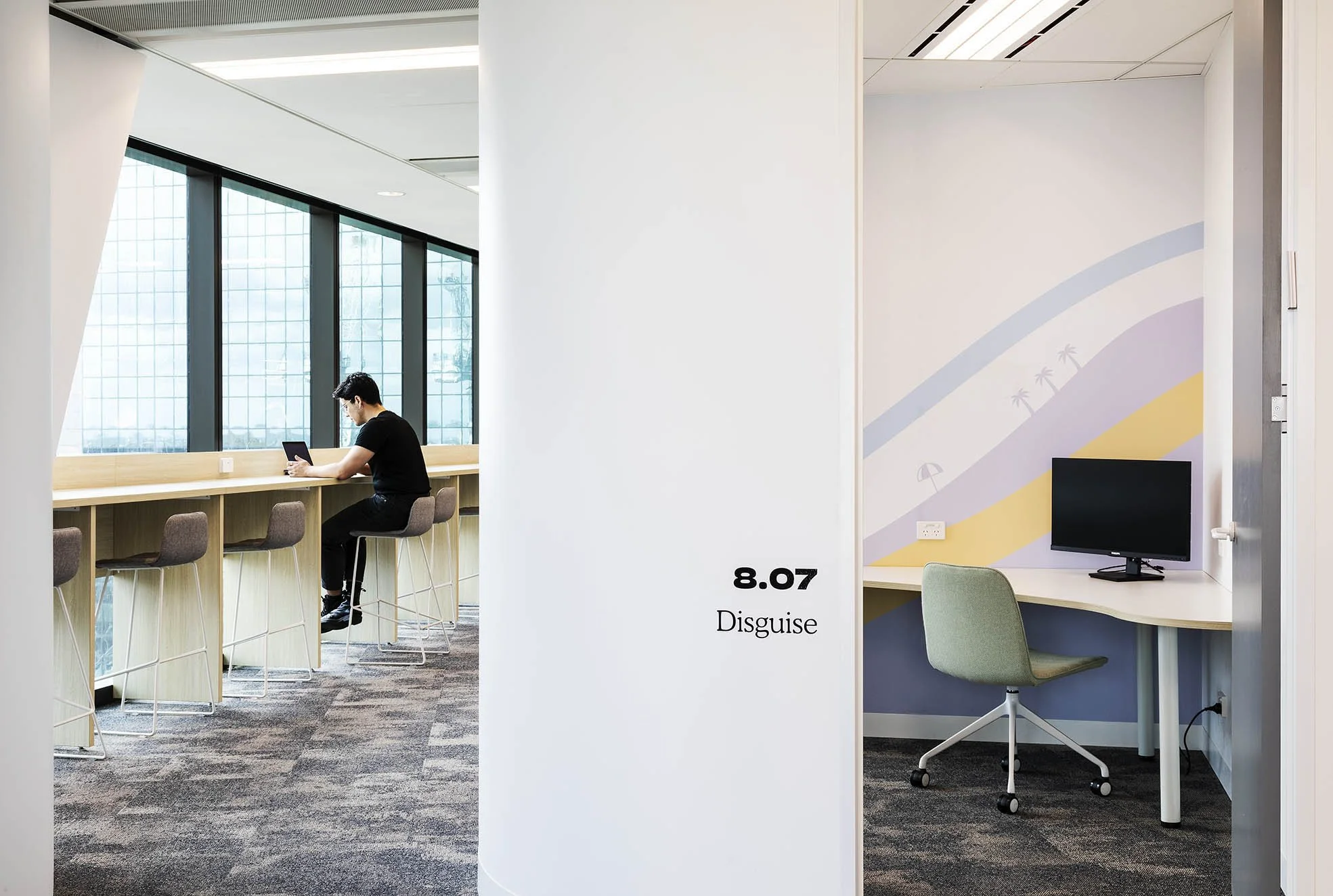 Interior of an office with two sections separated by a wall; on the left, a man sitting at a high table using a tablet near large windows, and on the right, a small work area with a computer monitor and a green swivel chair, with a colorful patterned wall and ceiling vents.