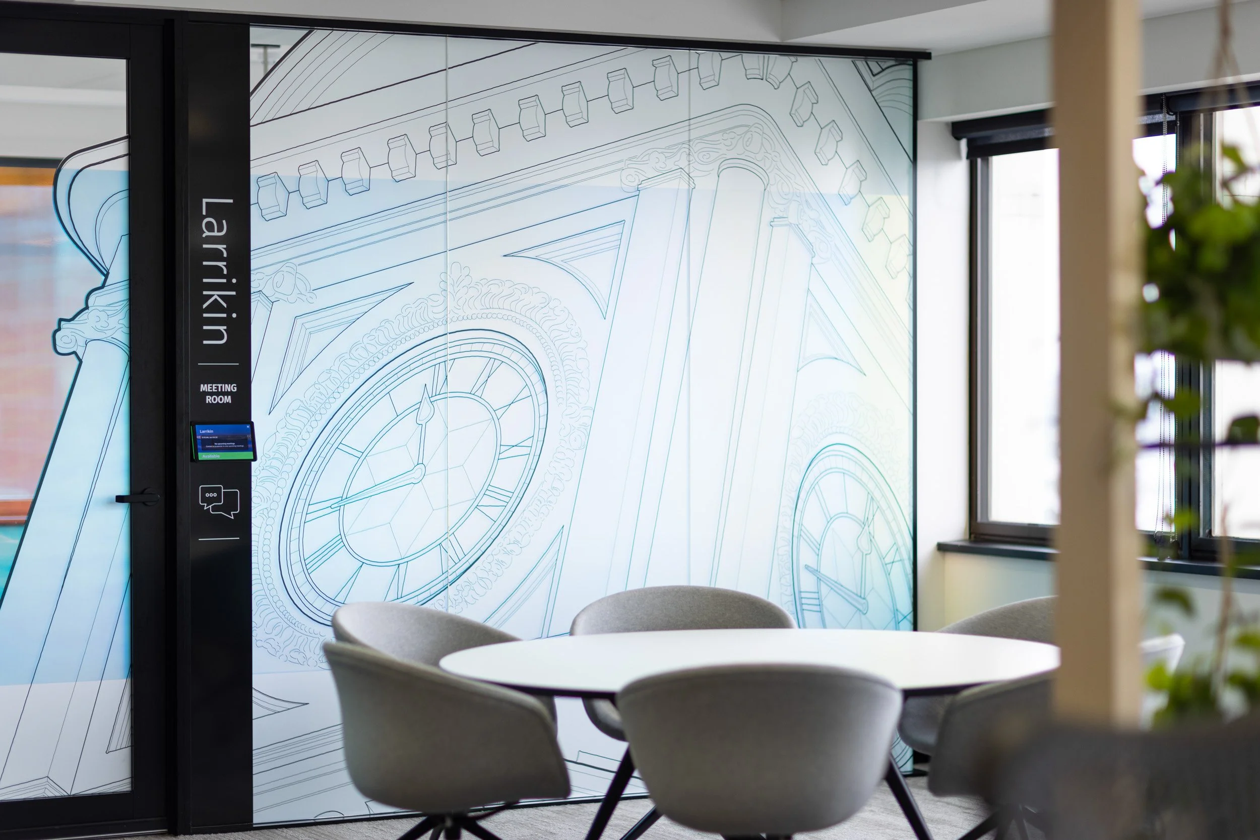 Interior of a modern office space with a meeting room. The room features a large glass wall with an outline drawing of a clock and architectural details. There is a small round table with four gray chairs surrounding it. A black vertical panel on the left displays the text 'Larrikin' and 'Meeting Room' below it.