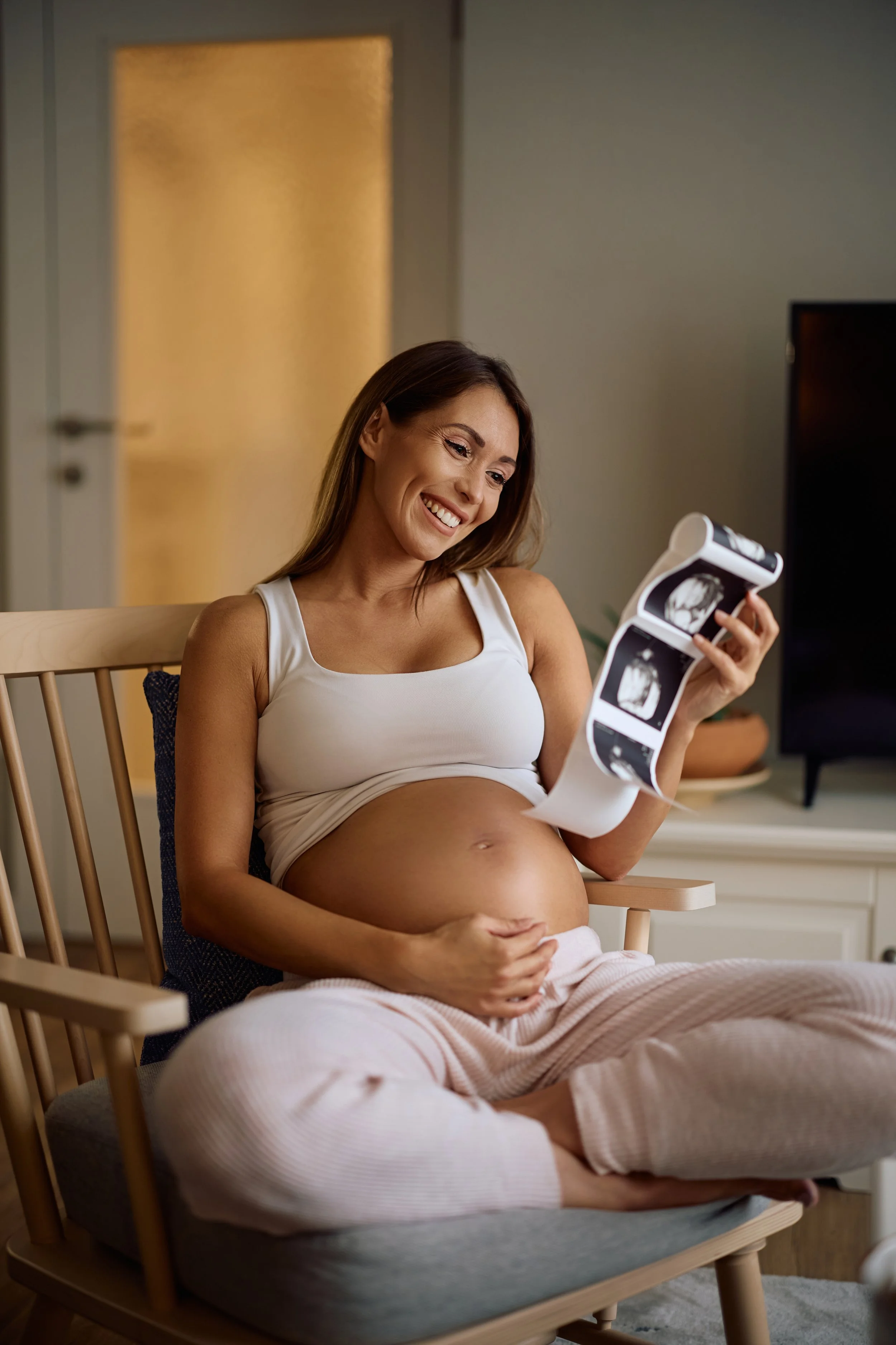 A pregnant woman sitting in a chair, smiling, and looking at ultrasound images she is holding.