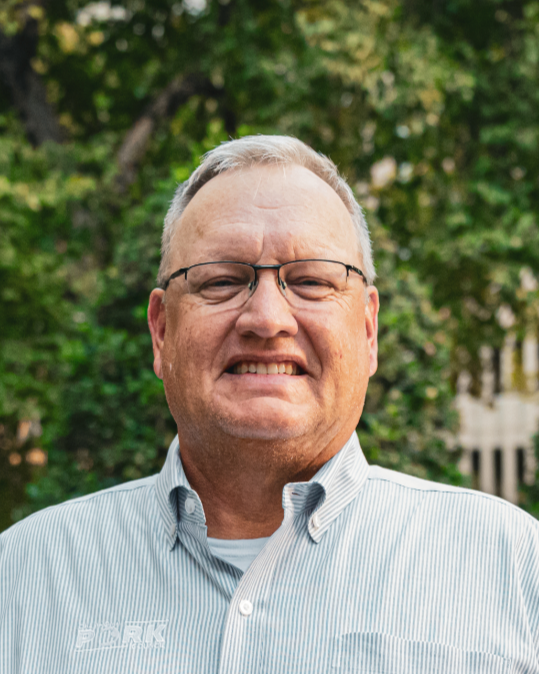 A smiling middle-aged man with gray hair and glasses, wearing a pink and white striped short-sleeve shirt, standing outdoors with a building and greenery in the background.