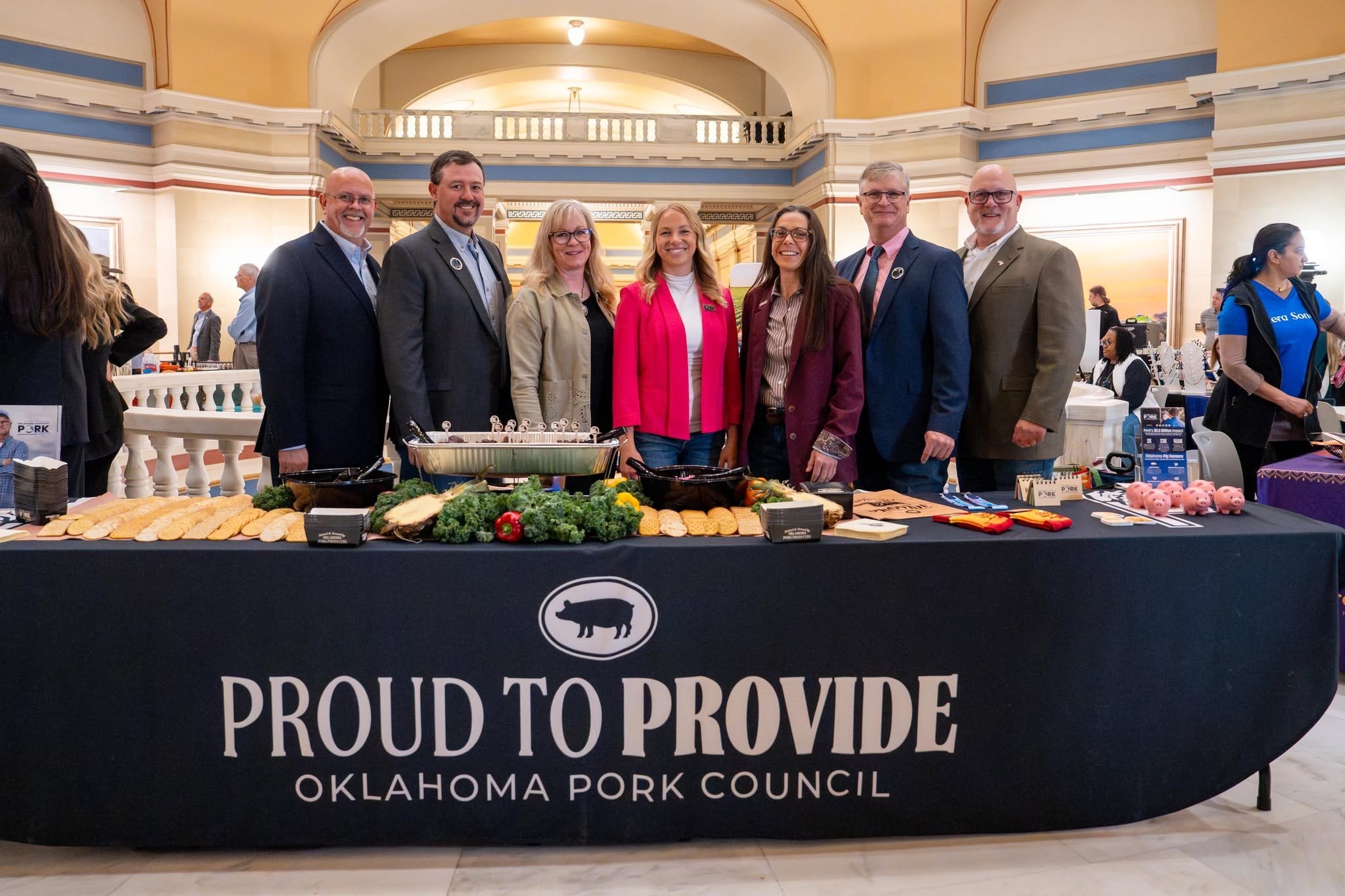 Group of seven people standing behind a table with food, at an event organized by the Oklahoma Pork Council. The table has a black tablecloth with the words 'Proud to Provide' printed on it, and there are various dishes and decorations displayed.