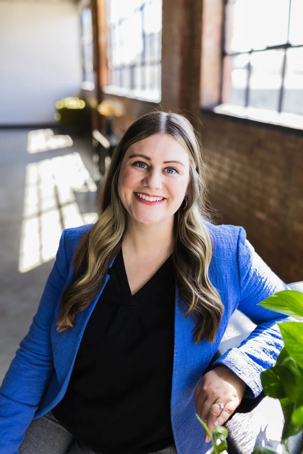A woman with long wavy hair, wearing a black top and a blue blazer, smiling and sitting near a large window in a bright, industrial-style room.