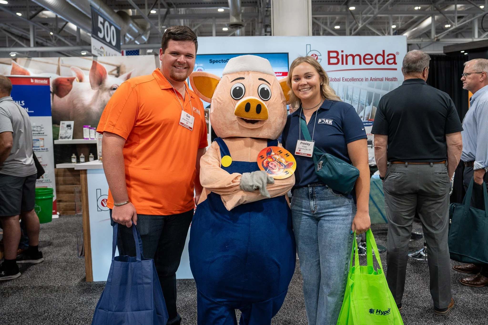 People at an exhibition, including two individuals smiling in front of a person in a pig costume. The background features a Bimeda booth and several other attendees.
