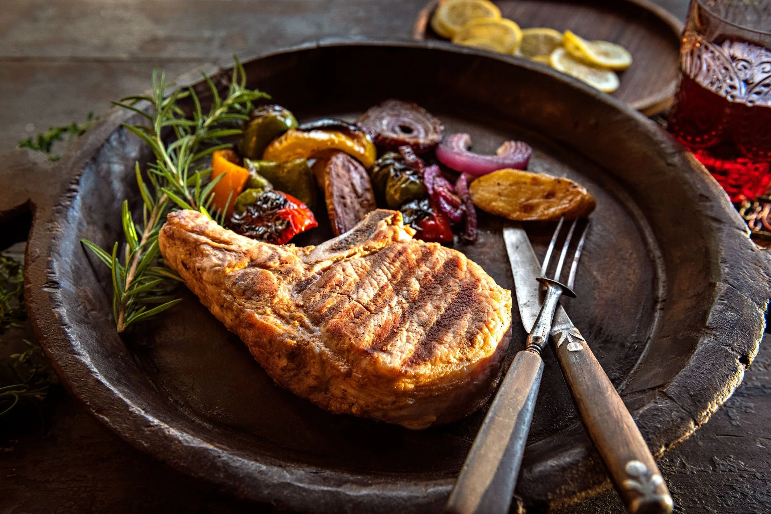 A rustic wooden tray with grilled fish, roasted vegetables, and sprigs of fresh herbs, with a fork and knife on the side.