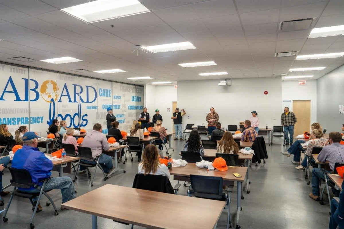 A group of people attending a meeting or presentation in a conference room, with some sitting at tables and others standing at the front. The room has a large wall display with the words "ABOARD cares" and related words in blue and yellow.