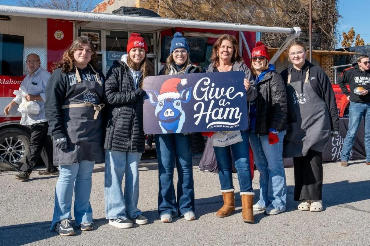 Group of people holding a sign that reads 'Give a Ham' in front of a food truck at an outdoor event during winter.