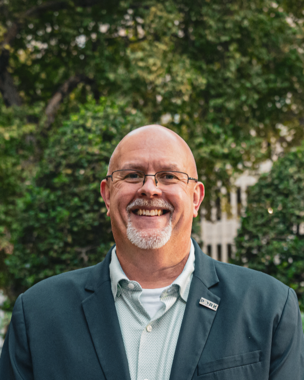 A smiling, bald man with glasses and a white goatee, wearing a blue blazer and a white shirt, standing outdoors in front of a blurred background with trees and buildings.