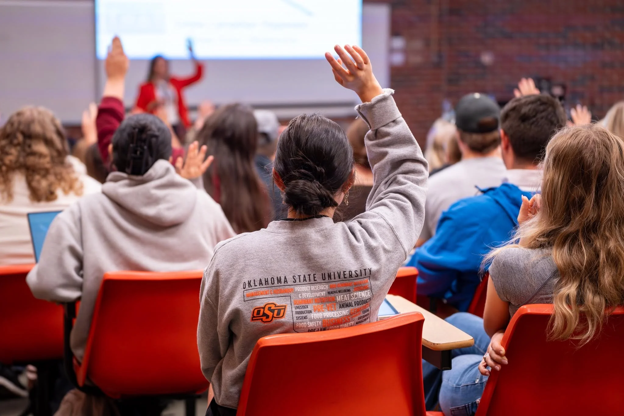 College classroom with students raising hands during lecture, focus on Oklahoma State University hoodie.