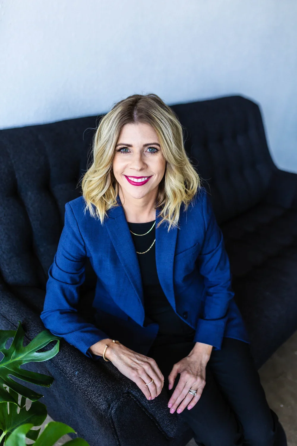 A woman with blonde, wavy hair and blue eyes, wearing a blue blazer, black top, and colorful jewelry, sitting on a dark sofa and smiling at the camera in a professional setting.
