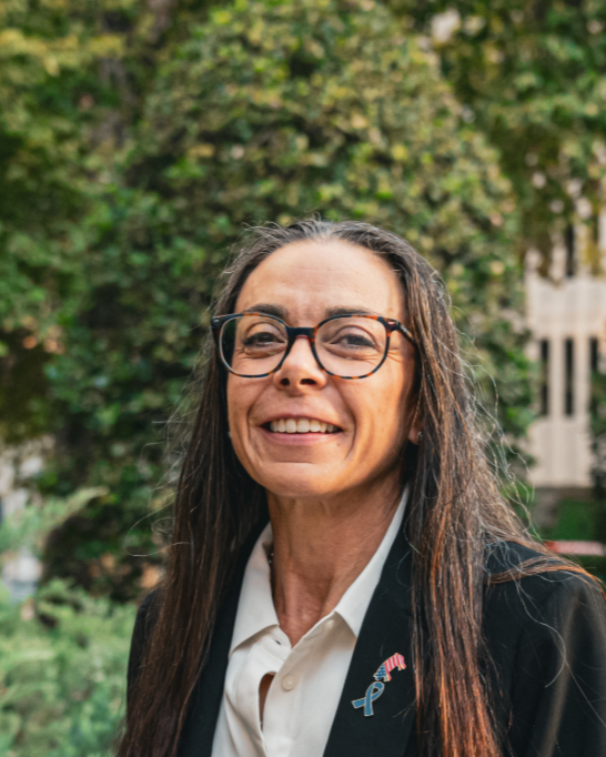 Smiling woman with long hair and glasses wearing a blazer outside with trees in the background.