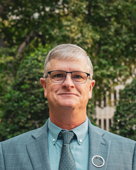 A middle-aged man with gray hair, glasses, in a blue suit and tie, standing outdoors near trees and a building.