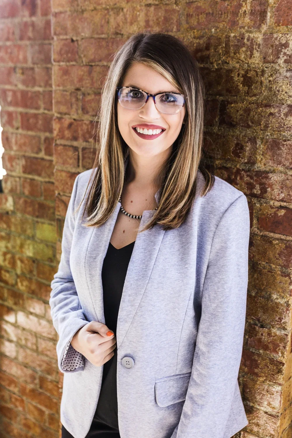 A woman with straight brown hair, wearing glasses, a gray blazer, and a black top, smiling and standing against a brick wall.