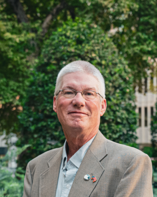 A mature man with gray hair and glasses wearing a beige blazer, maroon patterned shirt, and pins on his lapel standing outdoors.