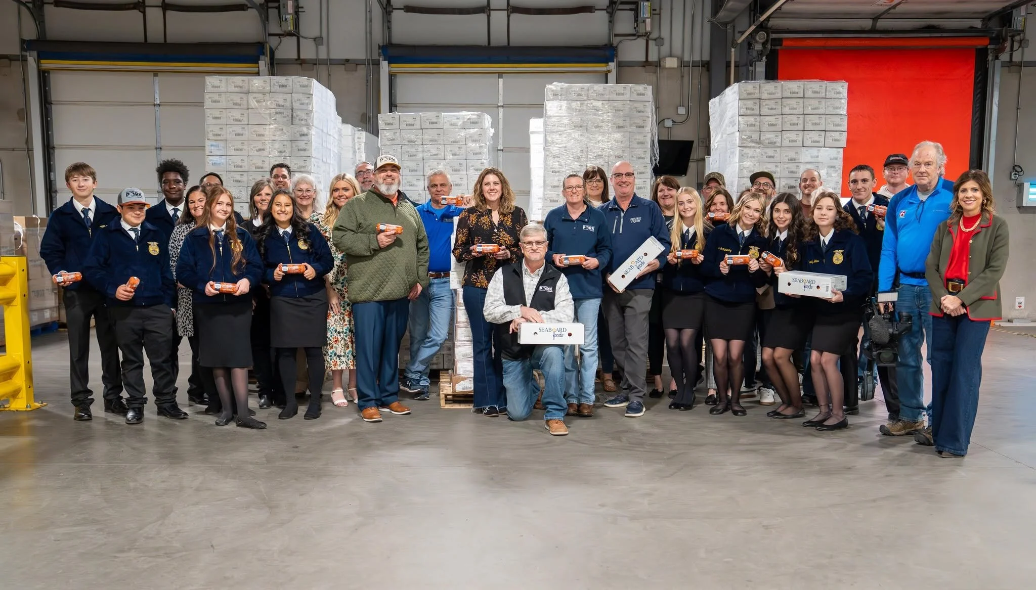 Group of people, including children in FFA jackets and adults, standing inside a warehouse with stacks of boxed goods behind them, some holding boxes of seafood.