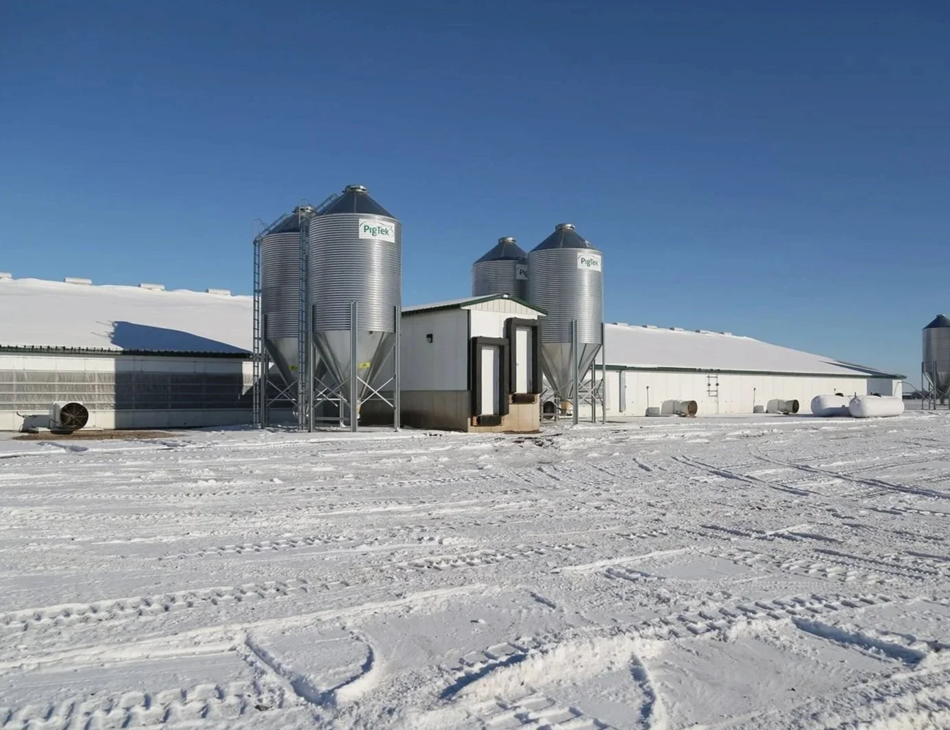 A snowy farm with large silos, a barn, and various equipment in bright daylight.