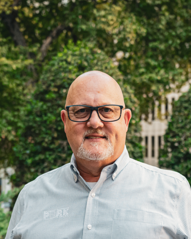 A middle-aged man with glasses, a smile, and a beard, wearing a light blue button-up shirt, standing outdoors with trees and a building in the background.