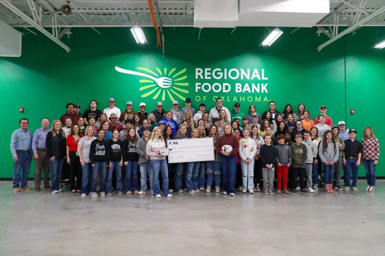 A large group of people, including children and adults, pose for a photo inside a facility for the Regional Food Bank of Oklahoma. Behind them is a bright green wall with a logo and the name of the organization. Some individuals hold a giant check and small food items.